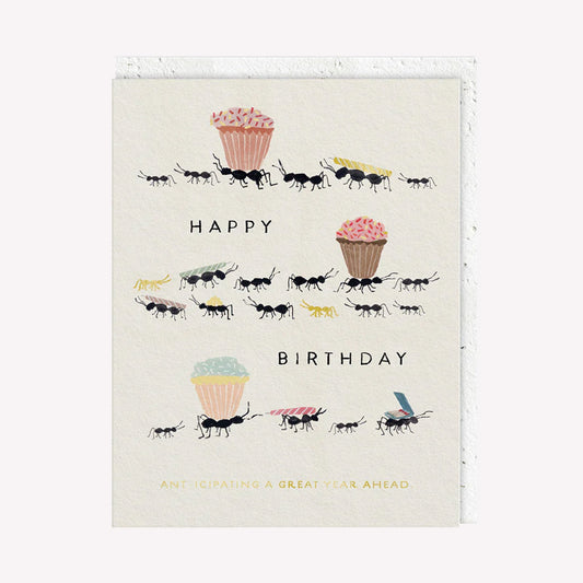 An illustrated greetings card by Seedlings, printed by Ohh Deer. This card features an illustration of ants carrying a birthday cake and candles, with text that reads “Happy Birthday / Ant-ipicating A Great Year Ahead”.