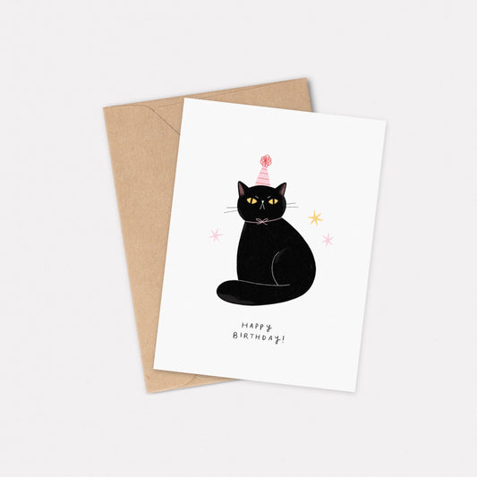 An A6 illustrated card featuring a sitting black cat wearing a pink striped party hat. Underneath the cat, text reads "Happy Birthday!". Behind the card is a kraft brown envelope.