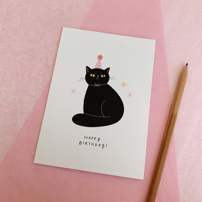 An A6 illustrated card featuring a sitting black cat wearing a pink striped party hat, with text underneath that reads 'Happy Birthday!'. The card lies on top of pink tissue paper and a pencil has been placed next to the card.