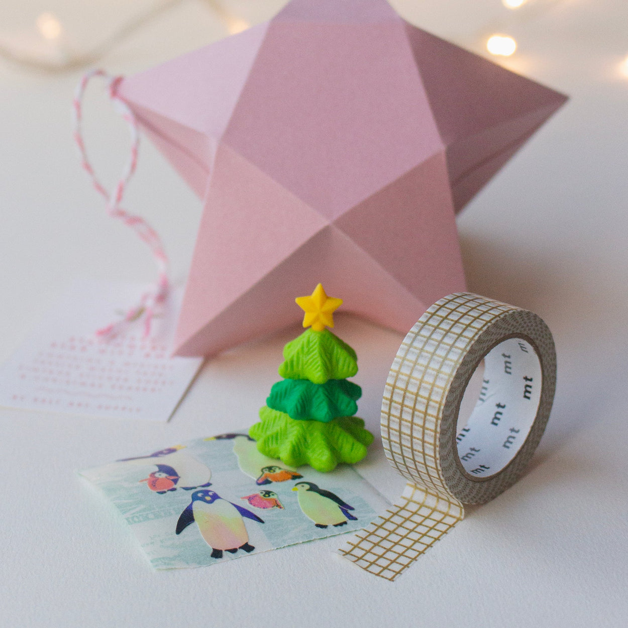Inside contents of Salt Art Supply's festive stationery star gift box, including a miniature tree shaped eraser, gold gridded washi tape and sheet of vintage penguin stickers. A pink origami star box can be seen in the background.