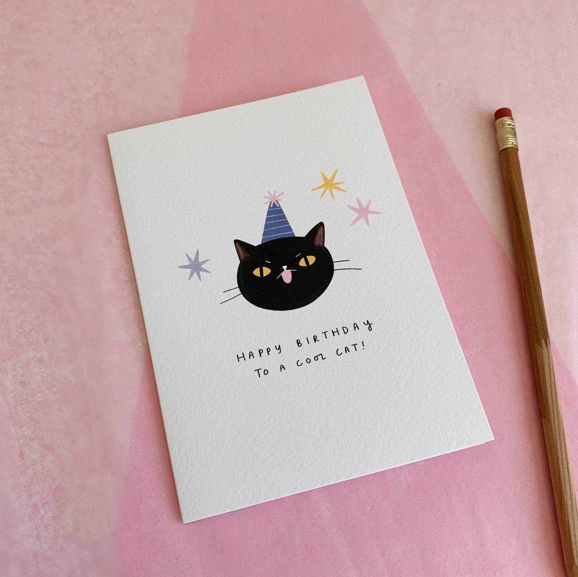 An A6 illustrated card featuring a black cat wearing a blue and pink striped party hat, with the text “Happy Birthday To A Cool Cat” underneath. Stars decorate around the cat face. The card lies on top of pink tissue paper and a pencil has been placed next to the card.