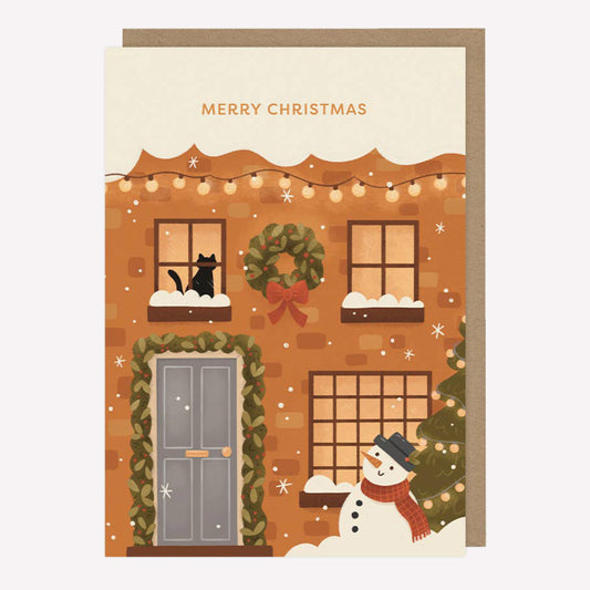 Illustrated Christmas greetings card featuring a snow covered house with a cat in the winodw and a snowman by the door with text reading "Merry Christmas". 