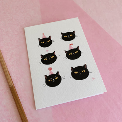 An A6 illustrated card featuring six black cat faces, some with pink striped party hats on. The card lies on top of pink tissue paper and a pencil has been placed next to the card.