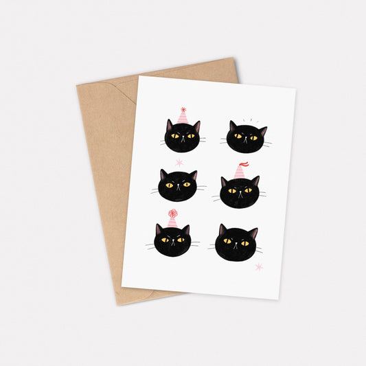 An A6 illustrated card featuring six black cat faces, some with pink striped party hats on. Behind the card is a kraft brown envelope.