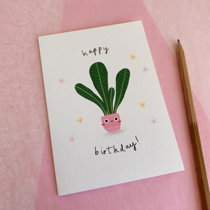 An A6 illustrated card featuring a house plant in a pink pot with a smiley face on. Around the house plant reads “Happy Birthday!”. The card lies on top of pink tissue paper and a pencil has been placed next to the card.