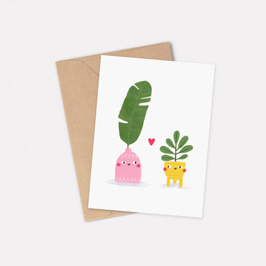 An A6 illustrated card featuring two brightly coloured house plants looking at each other, with a red heart between them. Behind the card is a kraft brown envelope.