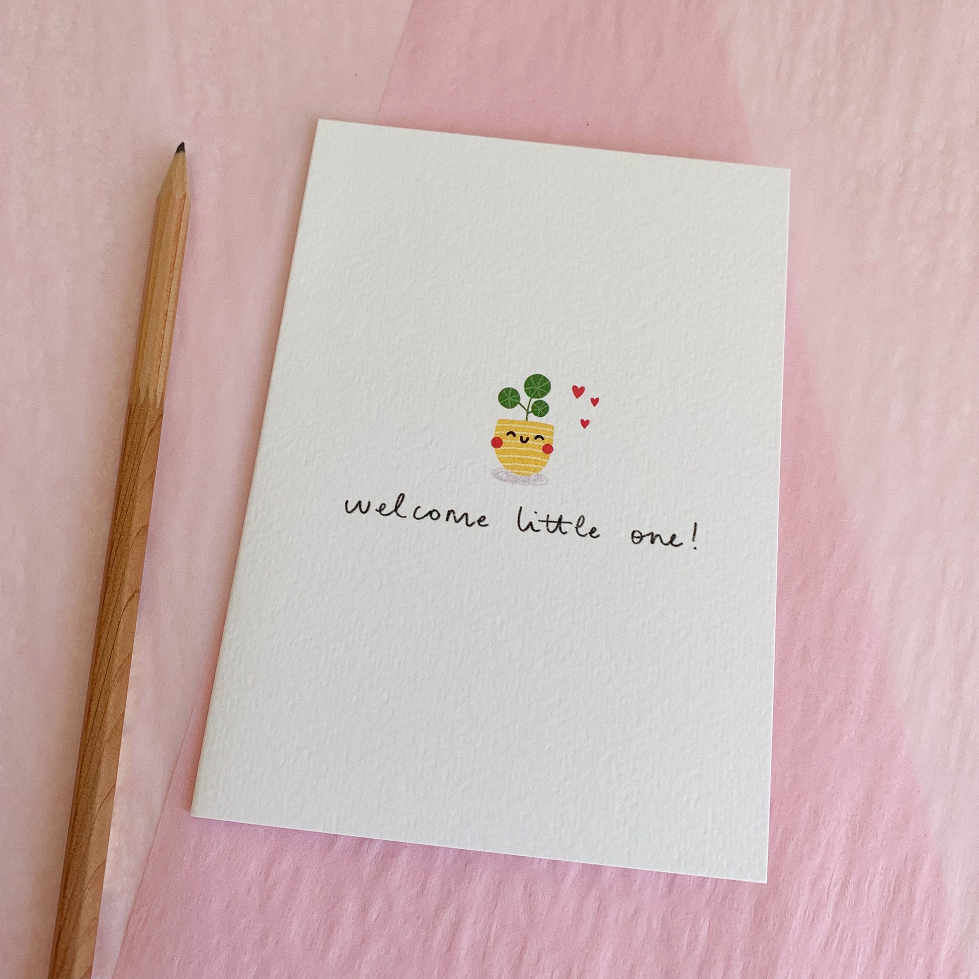 An A6 illustrated card featuring a small pilea plant in a yellow pot with a smiley face on it. Three hearts sit next to the plant, and it reads “Welcome Little One!” underneath. The card lies on top of pink tissue paper and a pencil has been placed next to the card.
