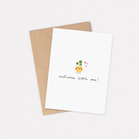 An A6 illustrated card featuring a small pilea plant in a yellow pot with a smiley face on it. Three hearts sit next to the plant, and it reads “Welcome Little One!” underneath. Behind the card is a kraft brown envelope.