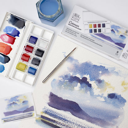 A lifestyle photo demonstrating the Cotman Watercolour Landscape Pocket set in use, with an open palette showing the 8 half pan paints included in this set, alongside a sea scape painting of a mountains and clouds over water to show the type of work this palette has been curated for. 