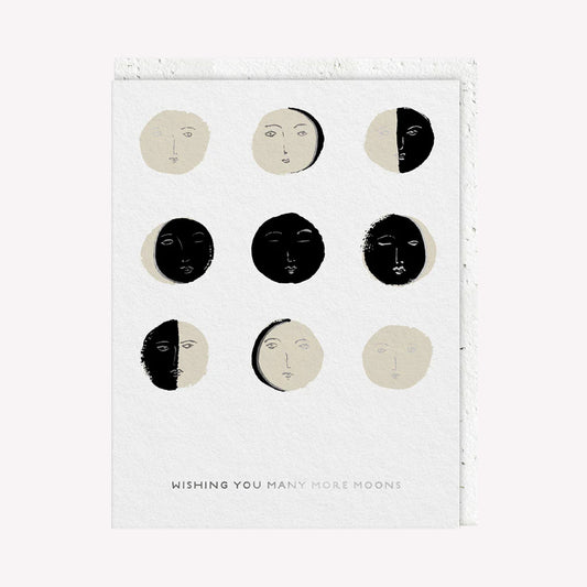 An illustrated greetings card by Seedlings, printed by Ohh Deer. This card features nine different moon faces, with text that reads “Wishing You Many More Moons”.