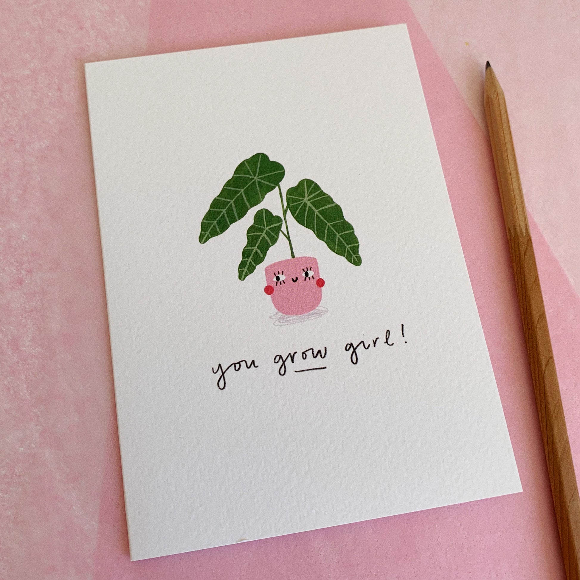An A6 illustrated card featuring a house plant in a pink pot with a smiley face on it. The text underneath reads “You Grow Girl!”. The card lies on top of pink tissue paper and a pencil has been placed next to the card.